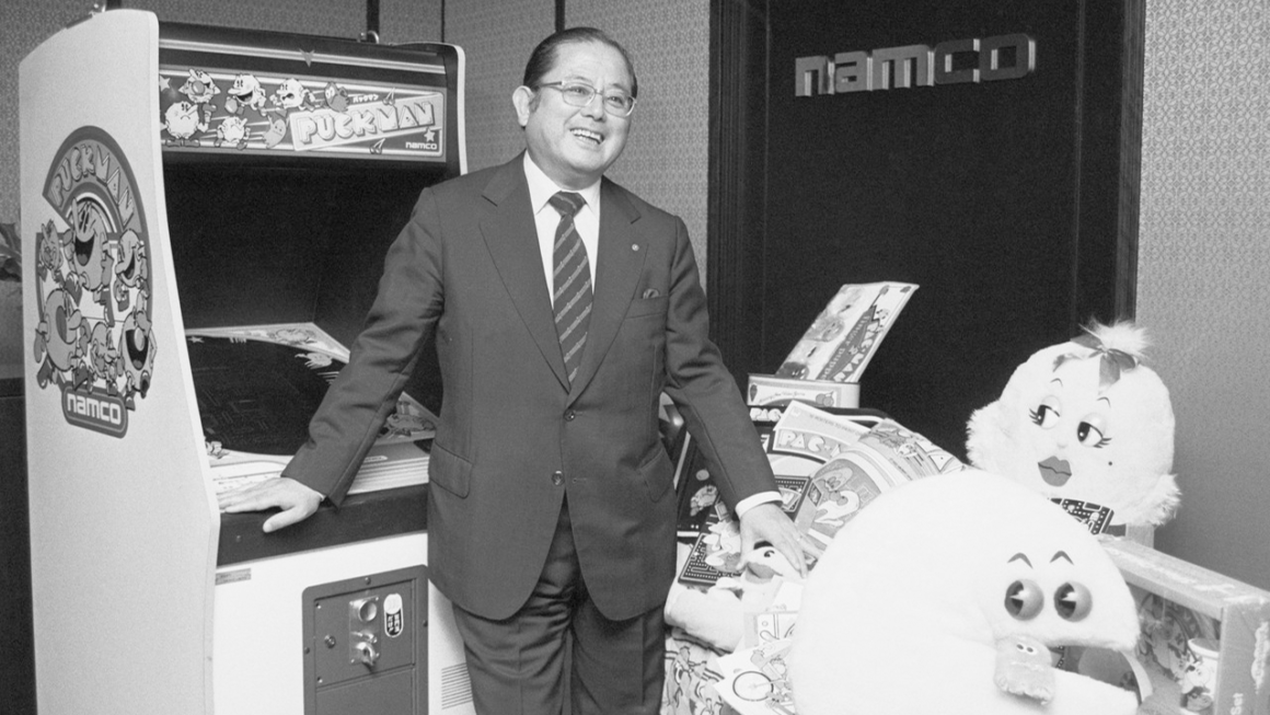 Namco's President and Founder Masaya Nakamura posing in front of a Pac Man arcade machine.